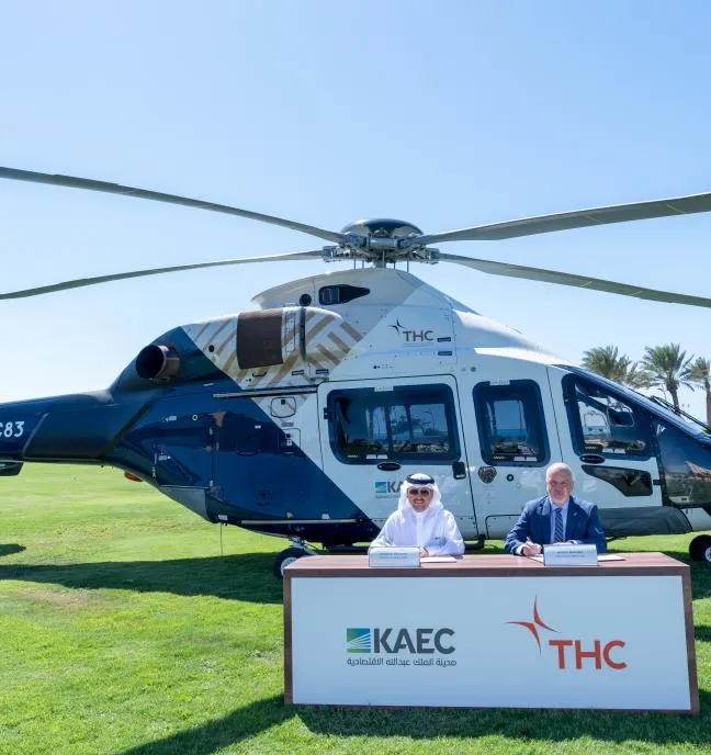 Man and woman seated at a table in front of a helicopter on a grassy field.